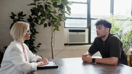 Especialista analizando un caso de psicología clínica en su consulta Una psicóloga clínica profesional con gafas y chaqueta blanca toma notas mientras mantiene una conversación terapéutica con un paciente joven en una oficina iluminada.