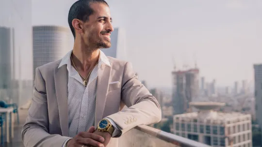 Profesional con habilidades de liderazgo frente a un paisaje urbano Hombre joven con traje claro y reloj de pulsera, apoyado en una barandilla de cristal con un paisaje urbano de edificios modernos al fondo, proyectando una actitud de liderazgo y visión.