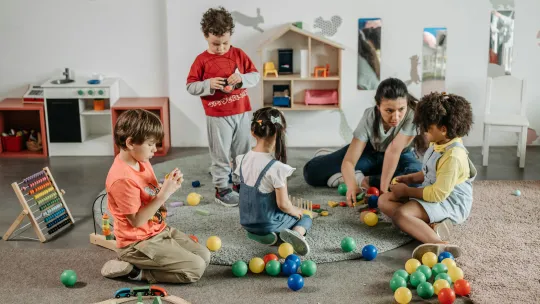 Educadora y niños compartiendo un momento de juego inclusivo en un aula infantil Niños jugando con pelotas y juguetes de madera junto a su profesora en un entorno de juego inclusivo para niños.